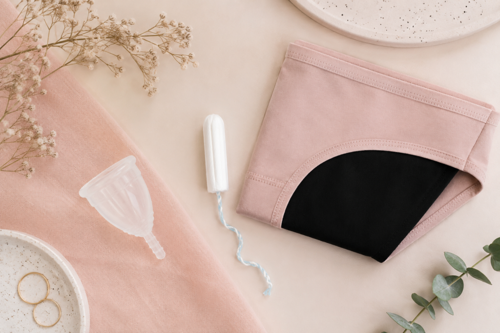 Uma composição flat lay minimalista e elegante em tons pastéis ou neutros (bege, rosa antigo, branco), exibindo de forma limpa um coletor menstrual de silicone transparente, um absorvente interno de algodão e uma calcinha absorvente moderna dobrada. O cenário deve transmitir leveza, Higiene Íntima na Menstruação e modernidade, afastando-se de conceitos puramente clínicos.]